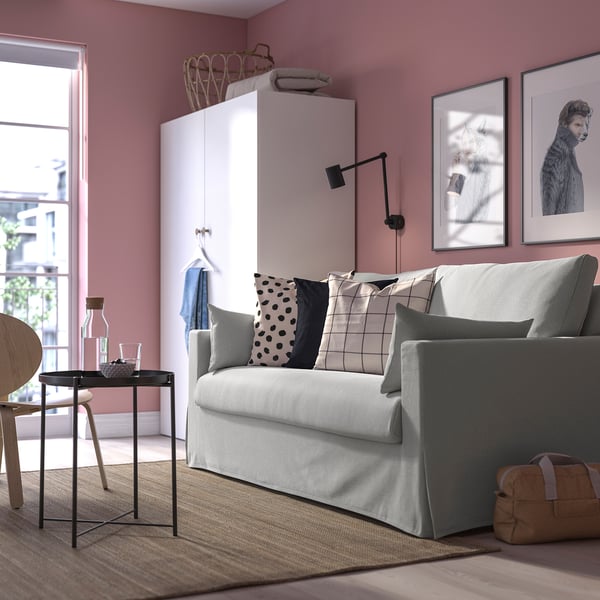 Cosy living room with grey sofa-bed, pink walls, patterned pillows, side table with vases, and white cabinet.