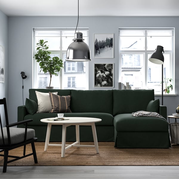 Modern living room with large green l-shaped sofa, white coffee table, and pendant lamp. Large windows and artwork on walls.