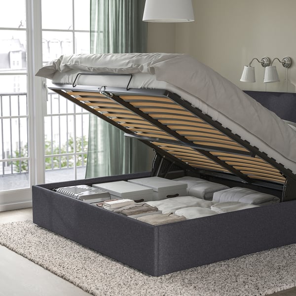 Bed with lifted storage compartment, showing organised blankets and pillows inside. Grey upholstered frame with slatted headboard.
