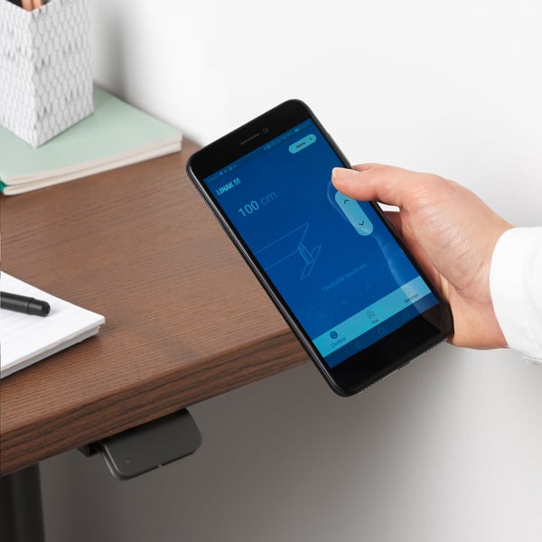 Person adjusts desk height using phone app. Smart desk app visible on phone screen.