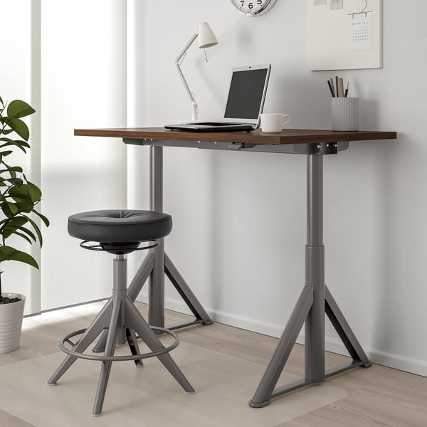 IDÅSEN brown/metal sit-stand desk with laptop, lamp, and stool. Large, fits room well, plant beside.