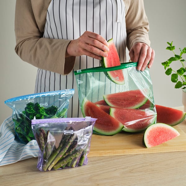 Person putting watermelon slice into reusable ISTAD bag. Table has more bags with broccoli and asparagus. Bags have sliders for easy sealing.