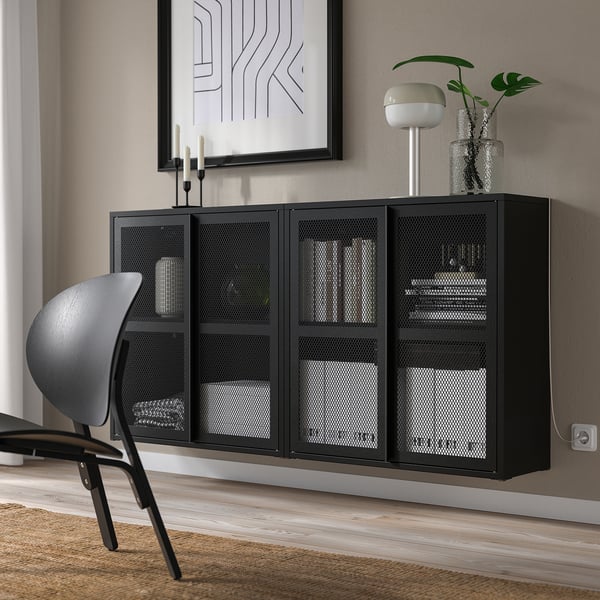 Black IVAR cabinet with mesh doors securely stores items on wall, between chair and artwork, contrasting with small decor.