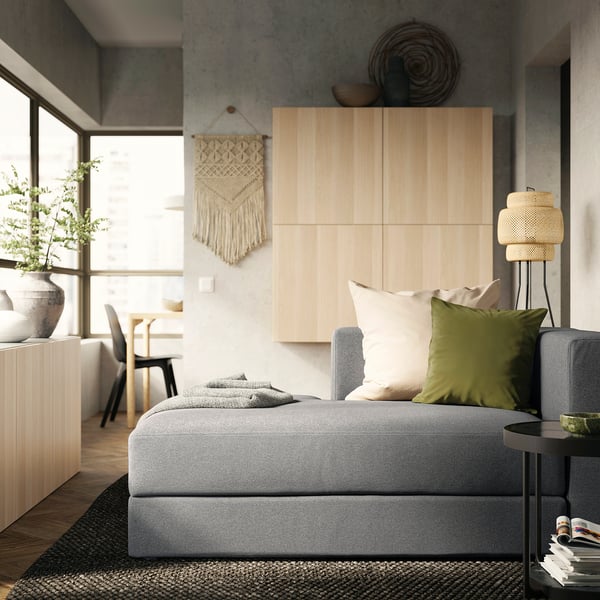 Modern living room with grey JÄTTEBO sofa, green pillows, woven wall art, wooden cabinet, and large window.
