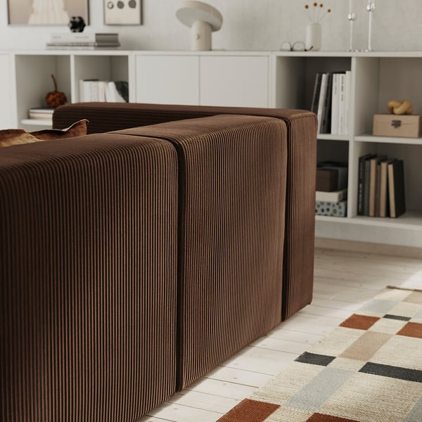 Brown modular sofa with cushions, positioned on a colourful rug, facing a white bookshelf with various items.