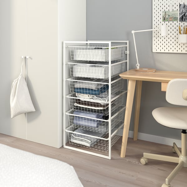 White JONAXEL storage unit with six drawers, placed beside a wooden desk and a chair. Great for organising items in any room.
