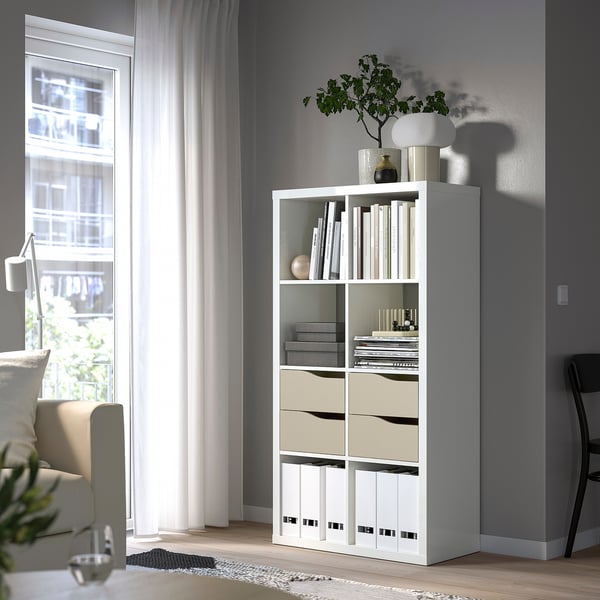 White KALLAX bookshelf near window, holding books, vases, and lamp. Cosy room with couch and rug. Larger than surrounding furniture.