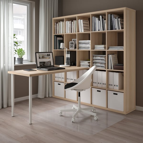 Modern home office with light wooden KALLAX bookshelf and desk. White swivel chair, laptop, and storage boxes.