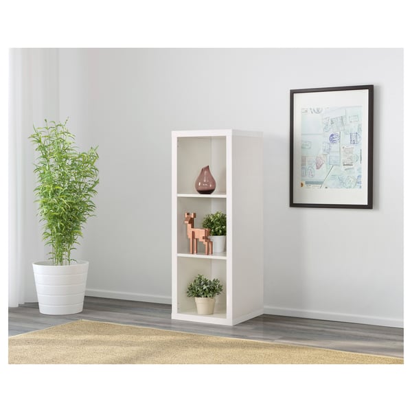 Grey walled room with KALLAX white shelving unit, holding vases and plant. Tall green plant beside it. Framed wall art nearby.