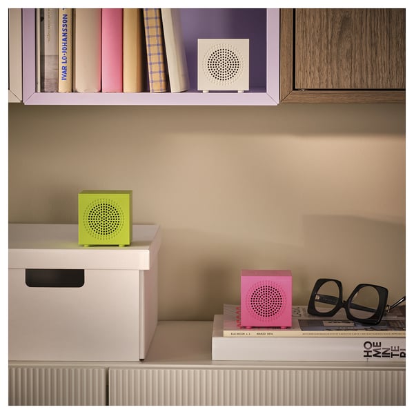 Three colourful cube speakers in green, pink, and white on shelves with books and a pair of glasses.