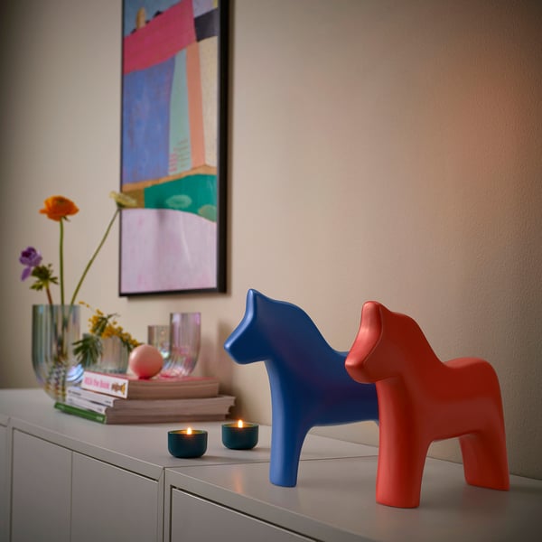 Two ceramic dala horses, one blue, one red, on a sideboard with books and flowers.
