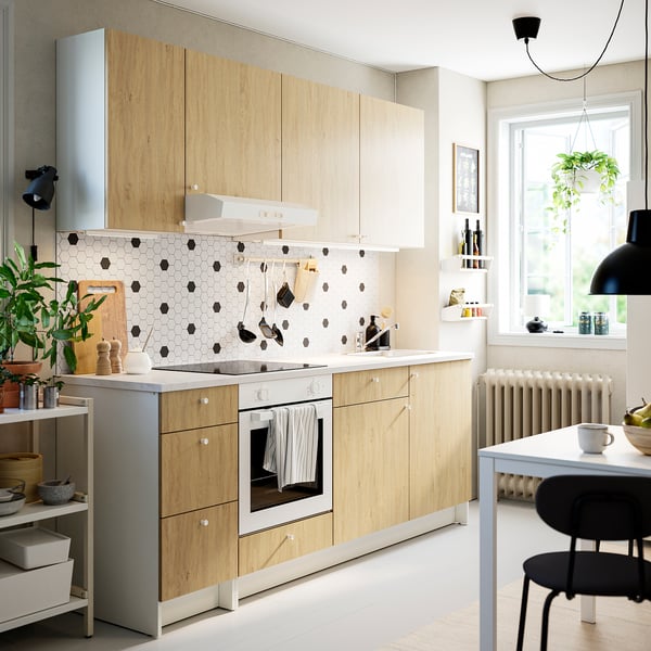 Modern kitchen with light wood cabinets, white backsplash, stainless oven, hanging plants, and black chairs around a dining table in a cosy area.