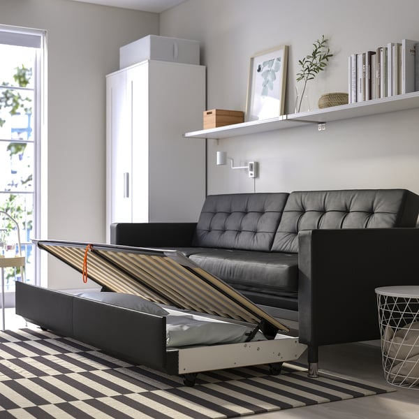 Modern living room with a black LANDSKRONA sofa-bed, partially opened showing wooden slats for sleeping comfort. White cabinet and striped rug nearby.