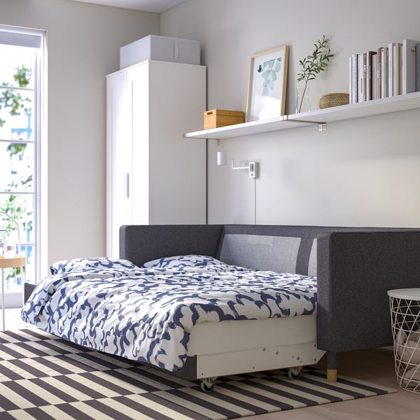 Modern bedroom with convertible sofa bed on wheels, white shelves with books and decor, large wardrobe, and striped rug under natural light.