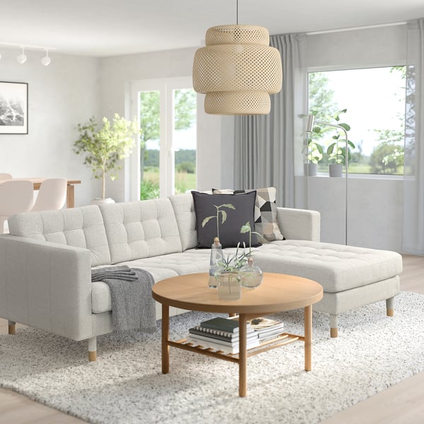 Modern living room: grey landskröna l-shaped sofa, natural wood coffee table, floral cushions, hanging textured lamp, large windows, indoor plants.
