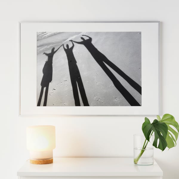 Black-and-white three peoples shadows holding hands, framed and displayed on a wall above a table with a lamp and plant.