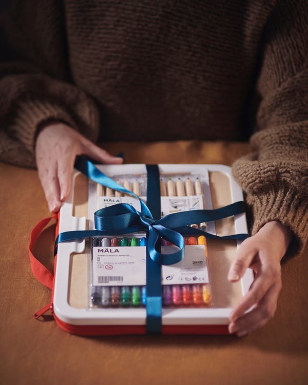 Person holding MÅLA drawing case, blue ribbon, art supplies visible, brown sweater.