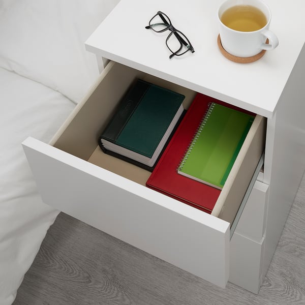 White MALM bedside table with open drawer, holding two books, glasses, and a tea cup. Safe for homes with kids.