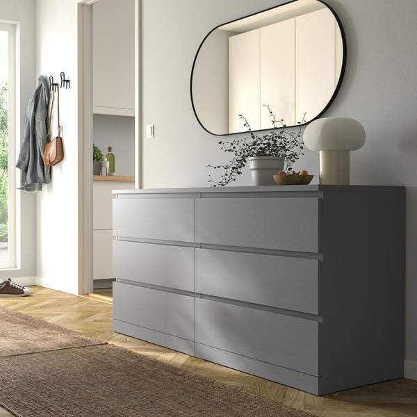 Modern bedroom with large grey MALM dresser, round mirror, lamp, & plants. Grey-stained wood, smooth drawers, wall anchored.