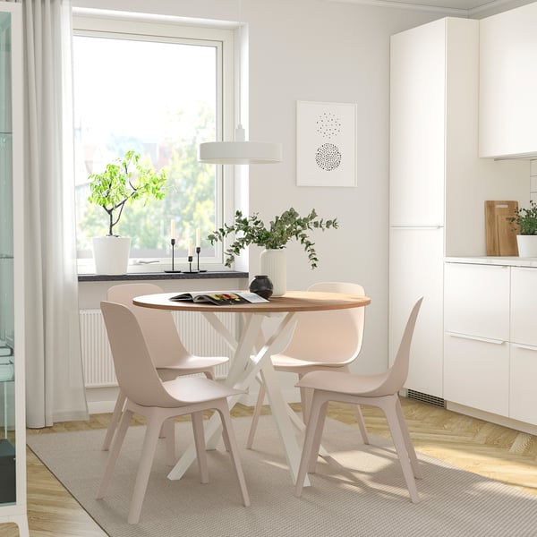 A modern dining area with a round MARIEDAMM table and light pink chairs, surrounded by a large window, potted plants, and minimalist decor.