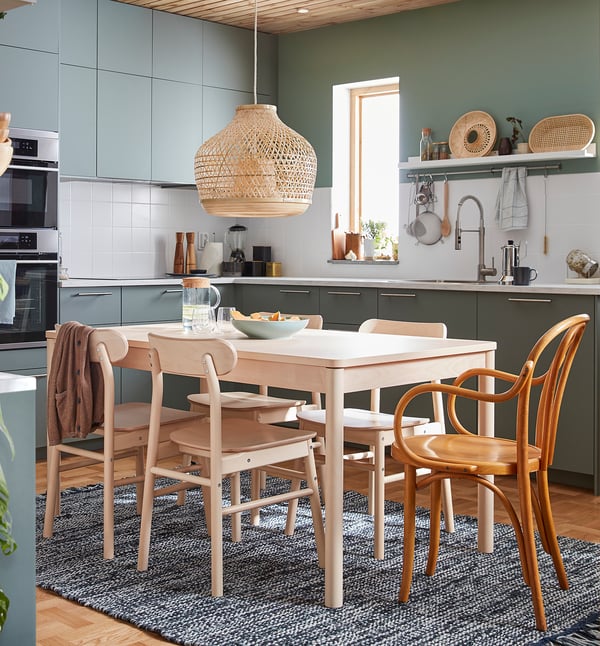 Modern kitchen with light wood dining set, green cabinets, hanging bamboo lamp, white countertops, and blue rug.