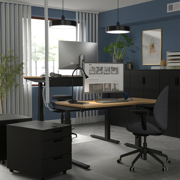 Modern home office with sit/stand desk MITTZON, black and wood, dual monitors, ergonomic chair, black cabinet, blue walls, large window, plants.