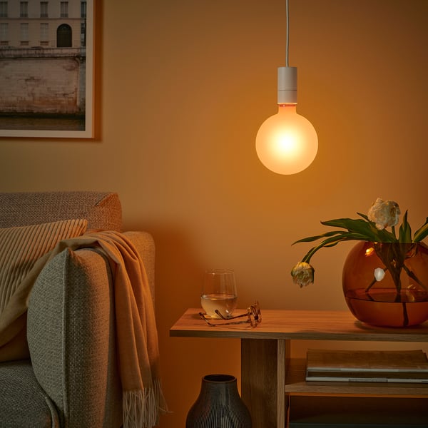A cosy living room with a MOLNART bulb casting a warm glow, near a couch with a blanket and side table with water, glasses, and flowers.