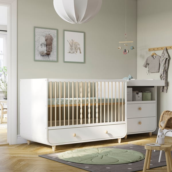 Cosy nursery with MYLLRA white crib, green wall art, soft rug, mobile, and changing table. Crib is large compared to other items.