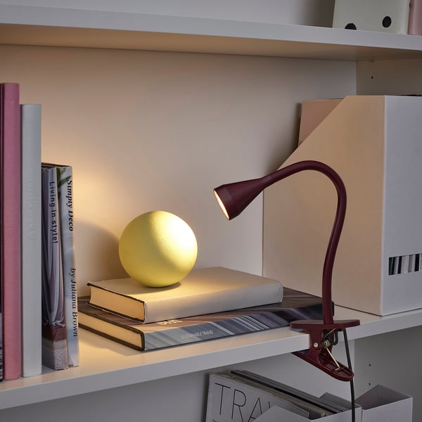 A burgundy NÄVLINGE clamp lamp on a shelf, illuminating a yellow sphere atop books. Slim and adjustable, it contrasts with larger books and boxes.