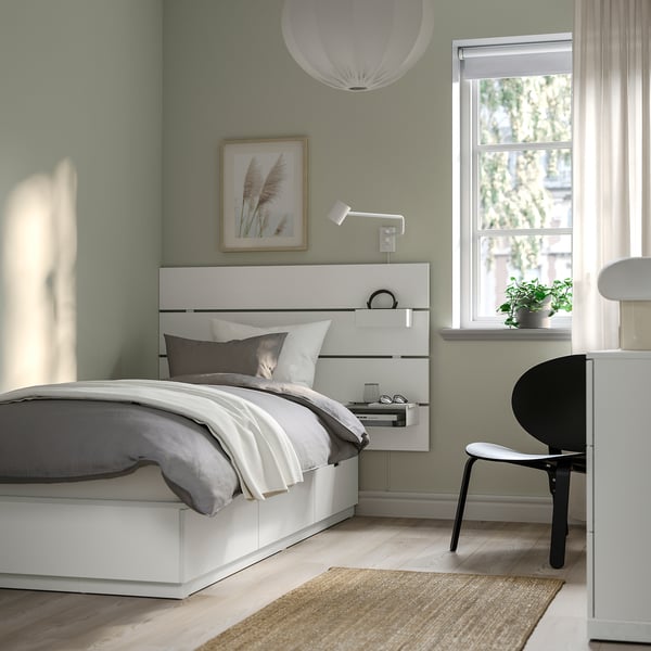 Minimalist bedroom with white NORDLI bed, grey linens, wall art, wall-mounted lamp, window with plant, black chair, rug, and light blue walls.