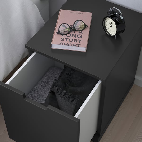 Bedside table with blanket, book, and clock beside white bed.