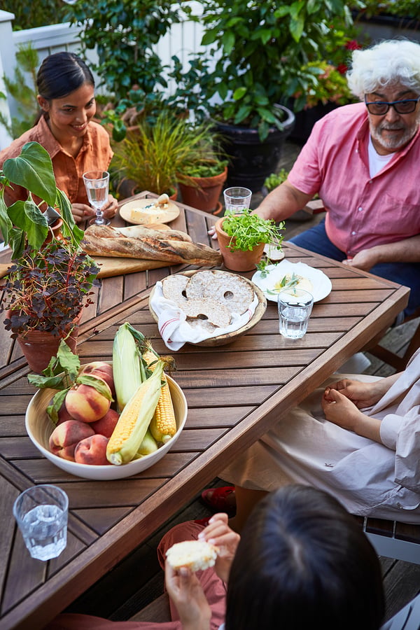 Three people enjoying a meal on a brown acacia wooden NORRMANSÖ table, surrounded by fresh food and greenery.