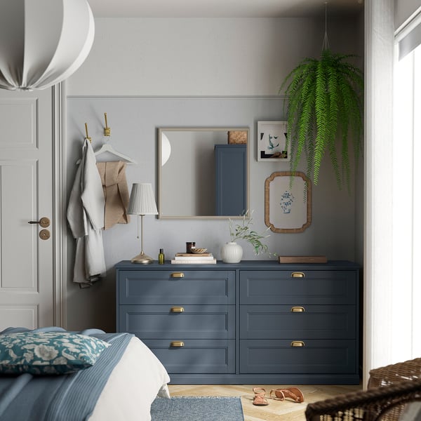 Grey bedroom with dresser, mirror, plants, and bed. Mirror is large and white-framed. Dresser is low with four drawers and gold handles.