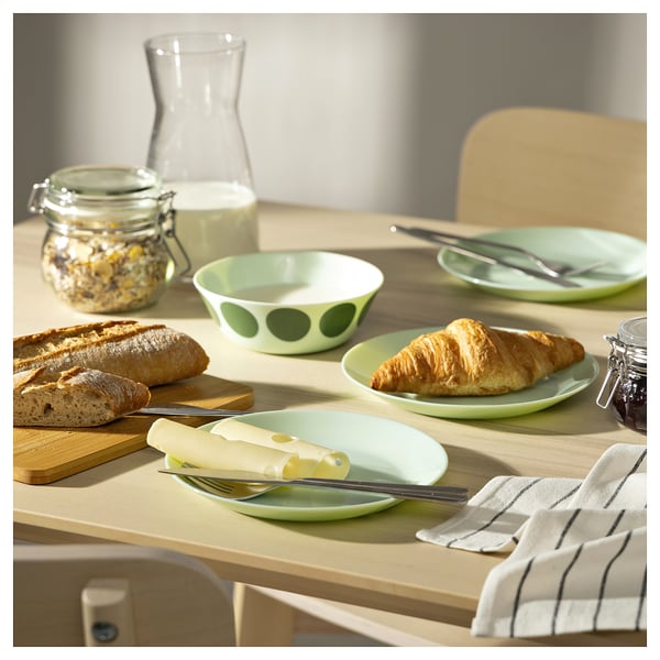 Green OFTAST tableware set on wooden table. Includes plates with croissants, cheese, bread, and utensils.