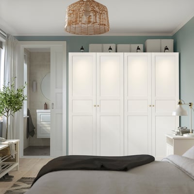 White PAX wardrobe with multiple doors/drawers against green wall, towering over bed with dark blanket and beige sheets.