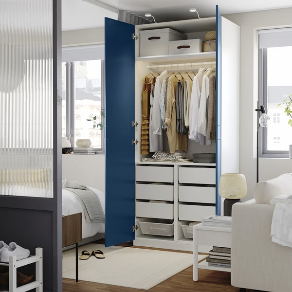 Open wardrobe displaying clothes, storage boxes, and drawers in a neat, organised bedroom with a window and chair.