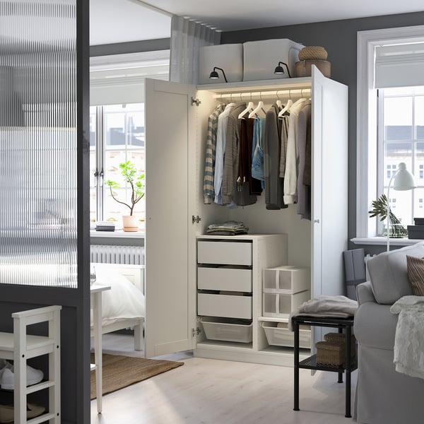 Modern white wardrobe with open space, lit inside, drawers, between bedroom and living room with chair, table, plant.