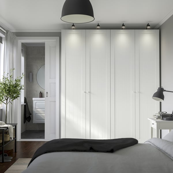 Modern bedroom with large white PAX closet, grey bed, plant, and desk lamp. Soft lighting highlights the wardrobes tall, sleek design.