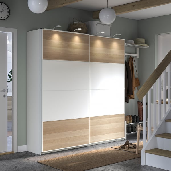 Large white and light wood PAX wardrobe with sliding doors, atop a rug, beside a staircase with hanging coats.