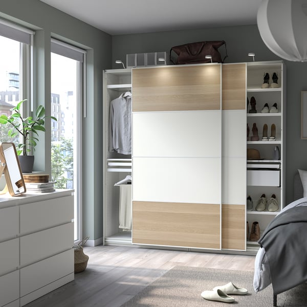 Modern bedroom with white and light wood PAX wardrobe, open to show clothes and shoes, next to a window.