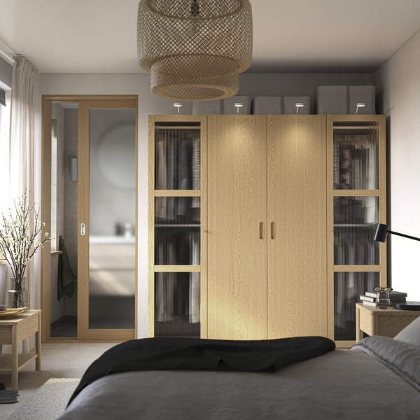 Serene bedroom with tall PAX wardrobe, glass panels, grey boxes, black bed, side lamp, warm natural light.