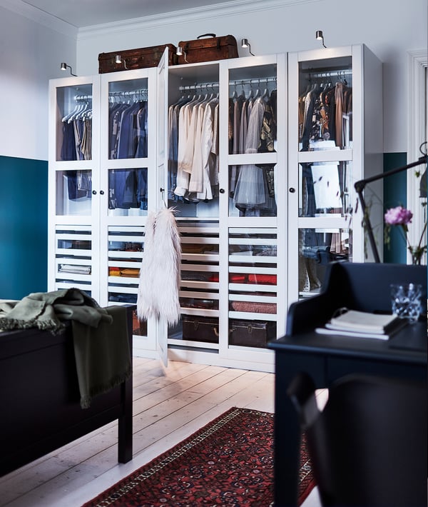 White PAX wardrobe with glass doors, clothes, and accessories. Wooden top, teal wall, patterned rug, organised drawers and shelves.