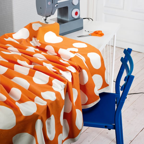 A sewing setup featuring orange fabric with white dots draped over a table, next to scissors, a sewing machine, and a blue chair, ready for crafting.