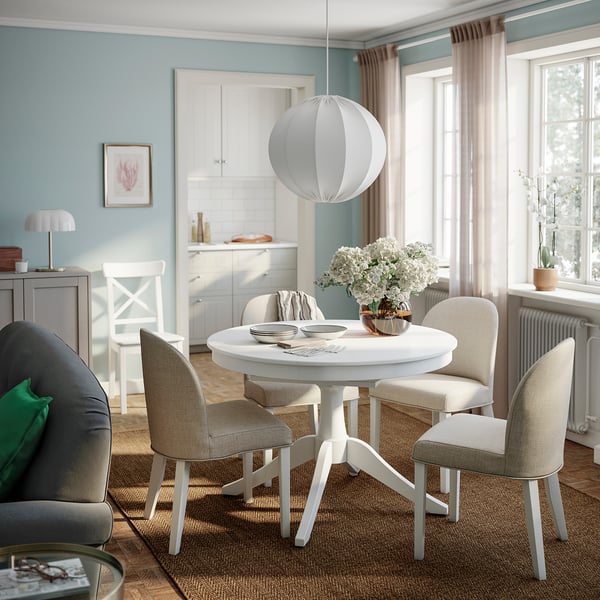 White extendable ROSENTORP table with beige chairs in cosy room, elegant pedestal base, central position, warm decor with light fixture and flowers.