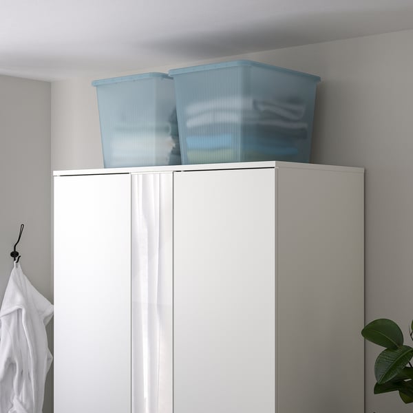 Two light blue, transparent plastic RYKTA boxes atop white wardrobe, larger than nearby white shirt.