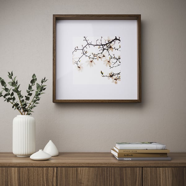 A framed blossoming branches hangs on a wall. Below it, a wooden shelf holds a white vase with greenery, two white candles, and stacks of books.