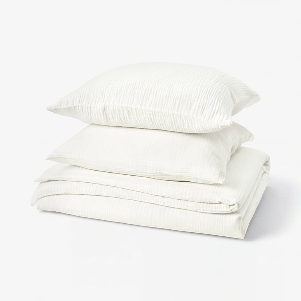 Stacked white bedding set, including folded duvet and pillows, featuring soft, textured muslin fabric.