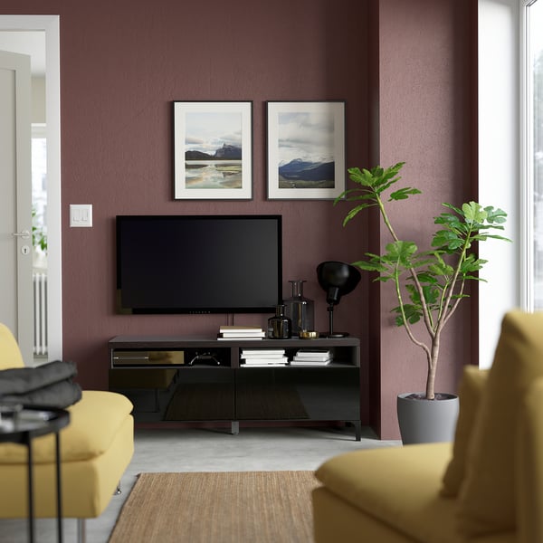 Modern living room with maroon walls, featuring a central tv on a glossy storage unit, yellow chairs, and mountain photos, balanced with greenery.