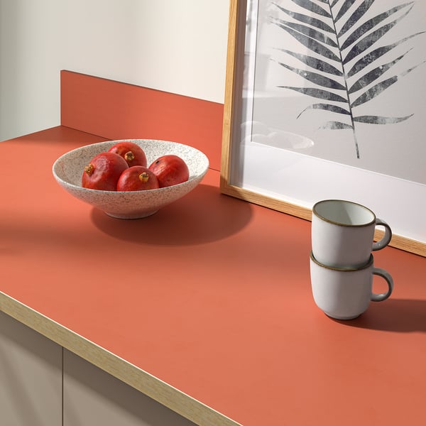 Bowl of red apples on a pink countertop, next to stacked white mugs, with a framed art print on a bright wall.