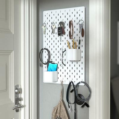 A white SKÅDIS pegboard organiser holds multiple items such as keys, pens, headphones, and a phone, offering a neat and accessible storage solution.